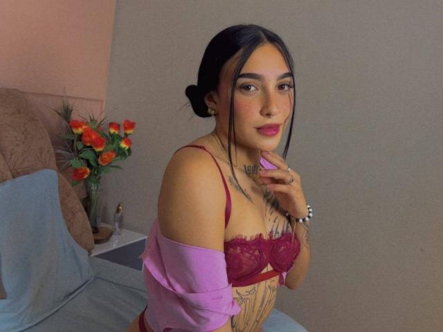 SophiieBlair with dark hair in a bun, wearing red lace bra, pink shirt off, tattoos on chest, pink lipstick, right hand near mouth, grey wall in background.