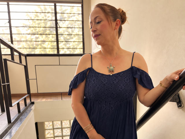 GianineLarose with bare shoulders, blue off-shoulder dress, medium tits, brown hair bun, necklace, standing by black staircase, right hand on rail, smiling, light skin, daytime.
