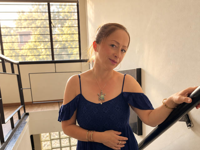 GianineLarose has light skin, brown hair in a bun, wearing a blue off-shoulder dress, holding a black railing, necklace with a green pendant, smiling slightly.