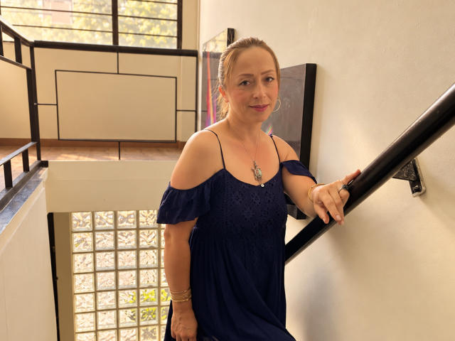 GianineLarose stands in a stairwell, wearing a blue off-shoulder dress, holding a black railing, with her hand on her ass, smiling at the camera.