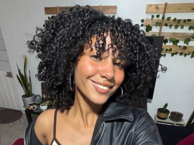 CinthiaWalker with curly black hair, smiling, wearing a black leather jacket, nose ring, and hoop earrings, showing her cute ass and shiny brown skin in a selfie.