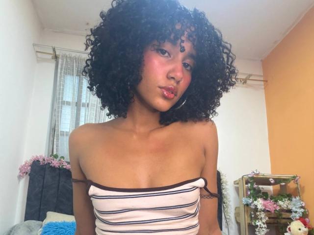 CinthiaWalker with curly black hair, pink blush, and a strapless white and black striped top, showing her small, perky tits. She has a cute, confident look.