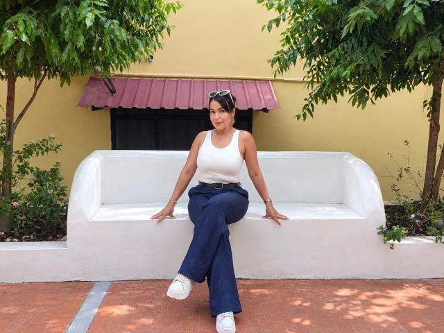 TamaraFoxy sitting on a white bench, wearing a white tank top, blue pants, and white sneakers, with sunglasses on her head, looking at the camera.