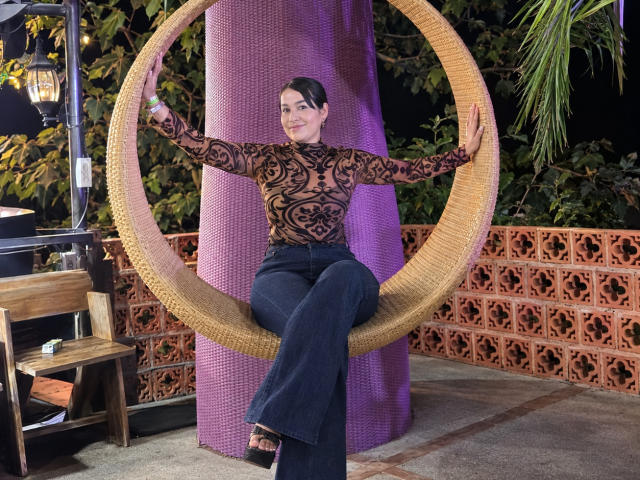 TamaraFoxy with small tits, wearing a sheer black patterned top, dark blue flared pants, sitting in a woven circular chair, arms spread, purple backdrop, night setting.