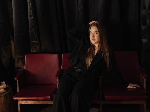 LauraHlot met lang bruin haar, een zwarte outfit en een riem, zittend op houten stoelen met rode kussens, met haar in haar handen, die lichtelijk opzij kijkt, op een donkere achtergrond..