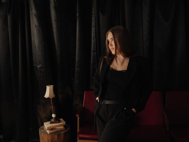 LauraHlot with long brown hair, black outfit, and belt, sitting on red chair, hands in pockets, looking to the side, next to wooden table with lamp. Dark background.