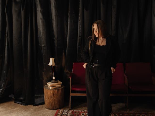 LauraHlot stands with hands in black pants, wearing a black top and belt, in front of a red couch, next to a wooden stool with a lamp.