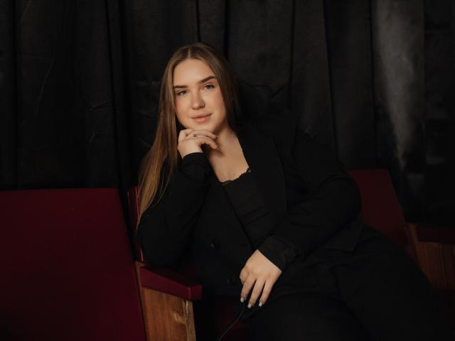 LauraHlot with long brown hair, black outfit, and subtle smile, resting her chin on her hand, sitting on a red chair, looking relaxed and confident.