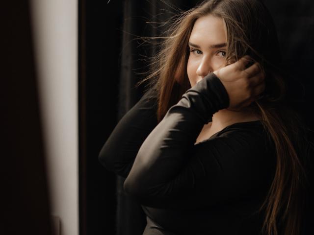 LauraHlot with long brown hair, wearing a black long-sleeve shirt, touching her hair with a slight smile, looking over her shoulder, in a dimly lit room.