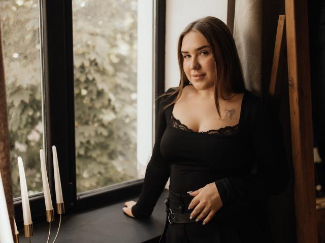 LauraHlot with medium tits, black lace-trimmed top, black belt, standing by window, brown hair, tattoo on right shoulder, holding window ledge, candles on left. Soft light.