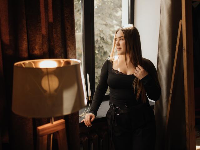 LauraHlot with long brown hair, black lace-trimmed top, and black skirt, standing by window, holding hair, under warm lamp light, looking outside. Boobs visible, curvy, sexy.