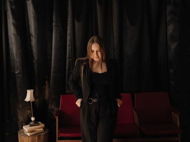 LauraHlot with long brown hair, black outfit, and belt, stands in front of red chairs, looking down with hands in pockets. Lamp on wooden table to the left.