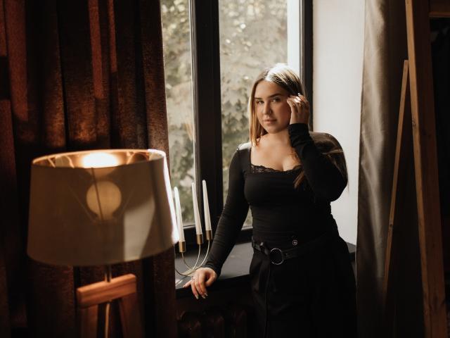 LauraHlot with blonde hair, black long-sleeve top, and black belt, standing by window, hand in hair, looking serious, table lamp with white shade in foreground.