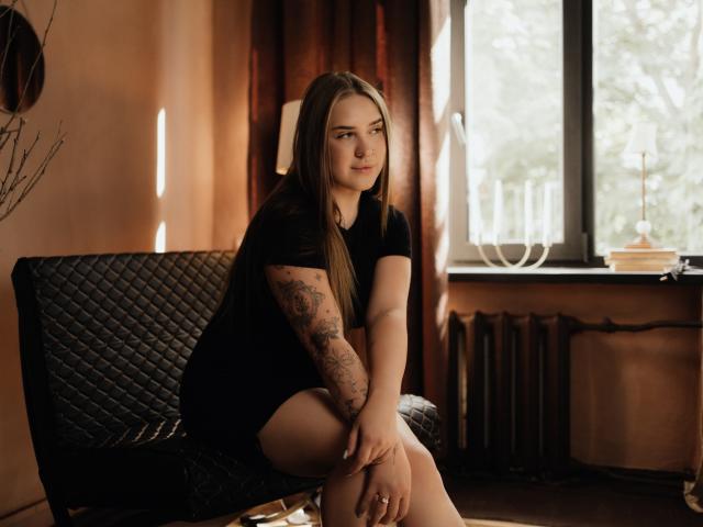 LauraHlot with long brown hair, black t-shirt, and tattooed arm, sitting on a quilted black chair, hands on knees, looking at camera, soft light, window in background.