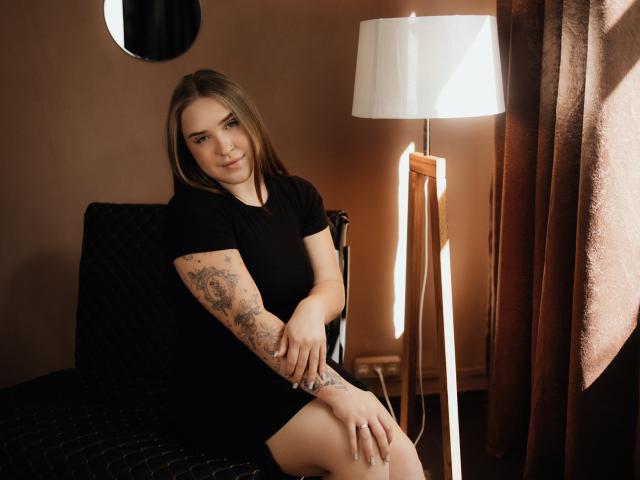 LauraHlot with tattoos on her right arm, wearing a black dress, sitting on a chair, smiling, hands on her knee, next to a white lamp, brown curtains, and a round mirror.