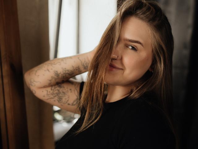 LauraHlot with blonde hair, nose ring, and tattoos on her arm, smiling slightly, wearing a black shirt, hand behind head, soft light.