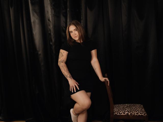 LauraHlot stands with one leg crossed, wearing a black dress, white socks, and showing tattooed arms. She leans on a chair with a leopard print seat.