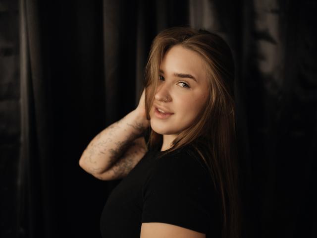 LauraHlot mit langen braunen Haaren, schwarzem Hemd, lächelnd, tätowiertem Arm, dunklem Hintergrund, Hand im Haar, süß, kleiner Nasenring, weiches Licht, leichtes Lächeln, entspannt, lässige Pose..