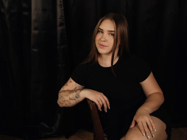 LauraHlot, con capelli castani, maglietta nera, braccio tatuato, seduta su una sedia, sorride leggermente, con una mano appoggiata sul ginocchio, sfondo con tenda nera, pavimento in legno, anello al dito, posa informale..