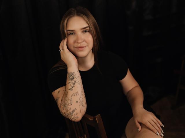 LauraHlot, con capelli castani, maglietta nera, tatuaggi sul braccio destro, con il mento appoggiato sulla mano, un leggero sorriso, seduta su una sedia di legno, sfondo scuro, che guarda verso la fotocamera..