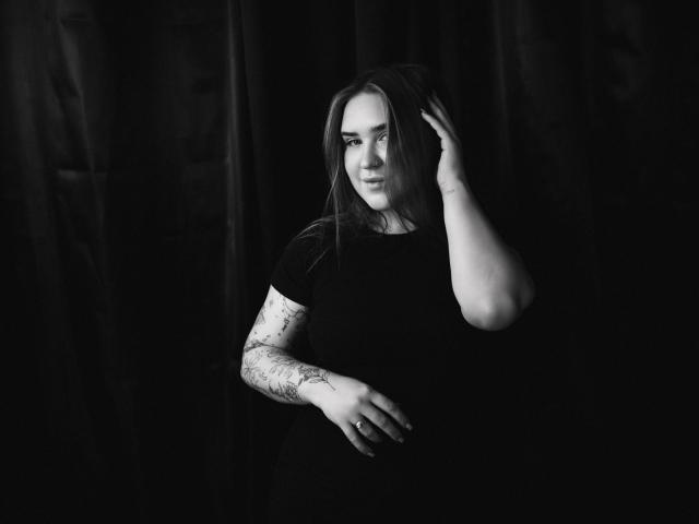 LauraHlot con capelli lunghi, camicia nera e braccio tatuato, mano tra i capelli, sfondo scuro, in bianco e nero, che guarda di lato con un leggero sorriso, primo piano medio, posa elegante..
