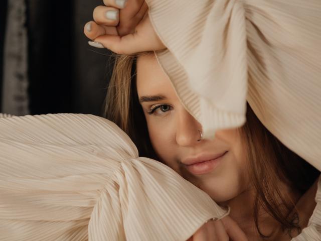 LauraHlot's face partially hidden by beige, pleated fabric, with her brown hair, green eyes, and a subtle smile. She's holding the fabric with white-painted nails.