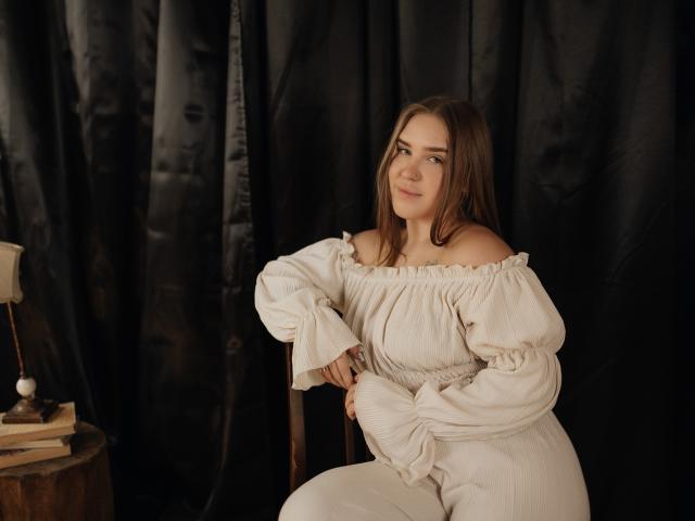 LauraHlot with brown hair, off-shoulder white dress, sitting on a chair, smiling, hands on lap, black curtain background, wooden table with lamp and books on left.