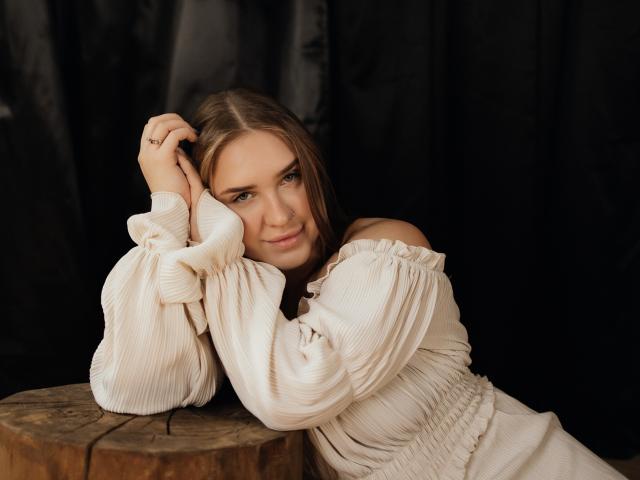 LauraHlot with long brown hair, off-shoulder white ribbed dress, leaning on wooden table, arms raised, subtle smile, blue eyes, ring on right hand, dark background.