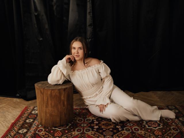 LauraHlot is leaning on a wooden stool, wearing a white off-shoulder dress, with her hand on her chin, on a patterned rug, looking relaxed and pretty.