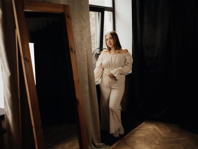LauraHlot stands near a window in off-shoulder white pants and top, looking to the side with a calm expression, her hands relaxed. Her tits are small, her ass is cute.
