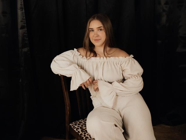 LauraHlot, with brown hair, sits on a chair wearing a white off-shoulder dress and pants, smiling slightly, her hand on her thigh, against a dark background.