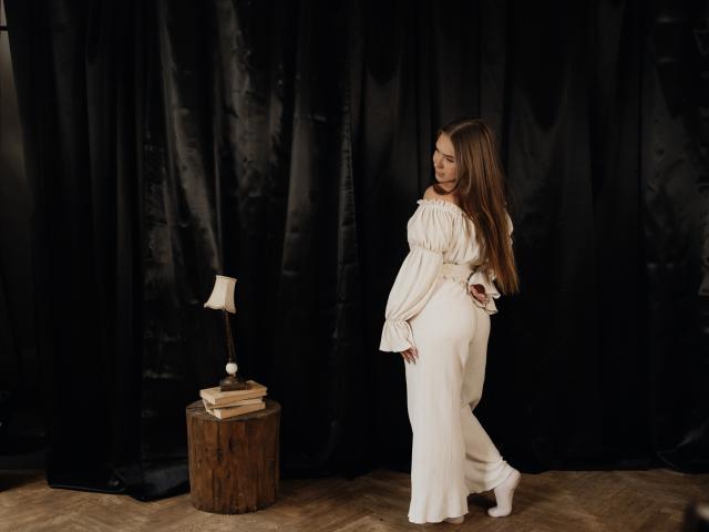 LauraHlot with long brown hair, white off-shoulder dress, and white high heels, standing by a wooden stool with a lamp and books, black curtain backdrop.