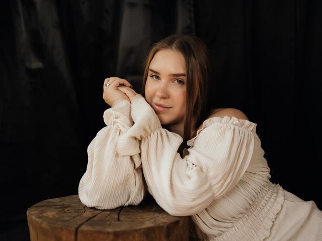 LauraHlot with brown hair, off-shoulder white ribbed shirt, resting arms on wooden table, subtle smile, soft light, dark background, casual, relaxed, cute, natural beauty, gentle expression.