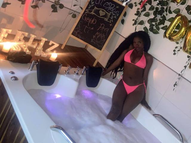 CatalinaSanA, com pele escura e cabelo cacheado, vestindo um biquíni rosa neon, sentada em uma banheira de hidromassagem cheia de bolhas, olhando para a câmera, com velas e plantas ao fundo..