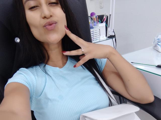 WettLady with dark hair, light blue ribbed shirt, pouty lips, and red nail polish, touching chin with finger, wearing a silver ring, in a black chair.