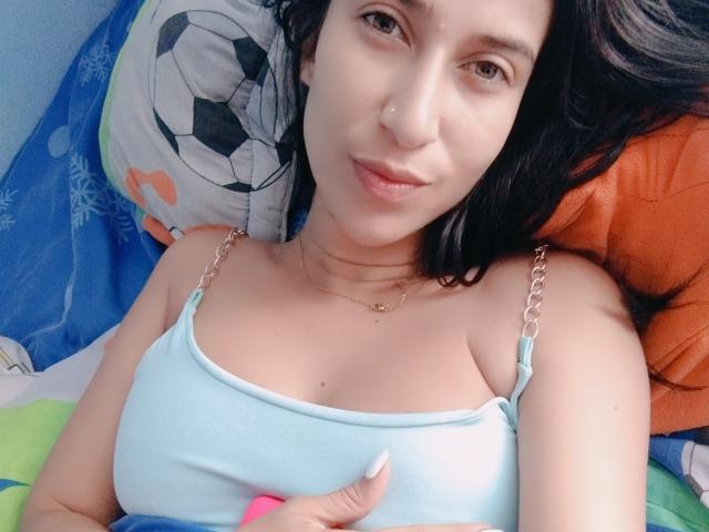 WettLady with light skin and black hair, wearing a light blue tank top with silver straps, lying on a bed, showing small tits, and touching her chest with pink nails.