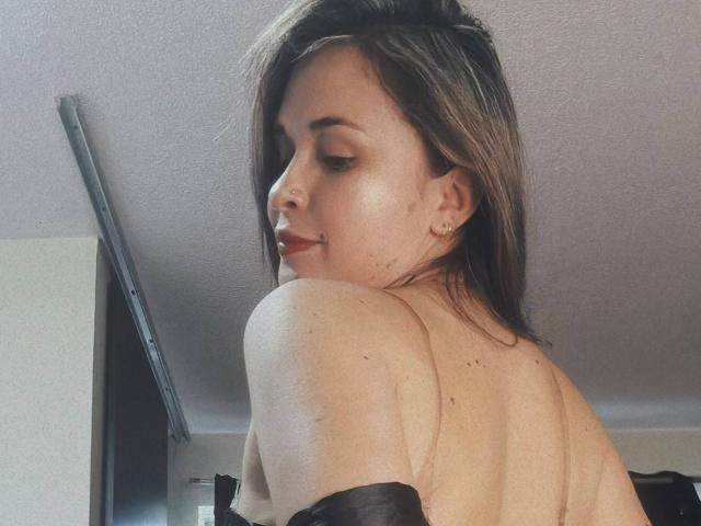 Wendy Schulz steht mit dem Rücken zur Kamera, wobei ihre nackten Schultern und der obere Rücken zu sehen sind. Sie hat langes, braunes Haar und trägt roten Lippenstift. Sie trägt einen schwarzen Träger..