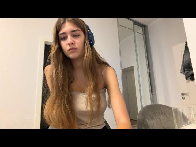 VerniaMiccio with long brown hair, blue headband, and a beige tank top, looking slightly bored, sitting in a white room with a mirror closet. Her tits are small.