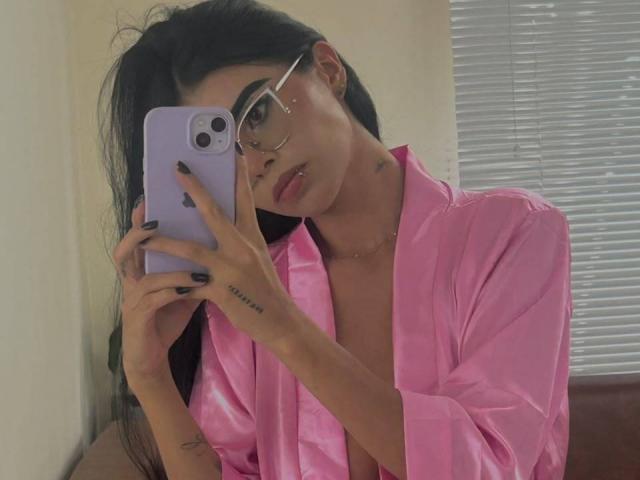 AmilyBella with long black hair, glasses, pink satin robe, taking a mirror selfie with a purple iPhone, showing tattoo on right hand, looking at phone with slightly parted lips.