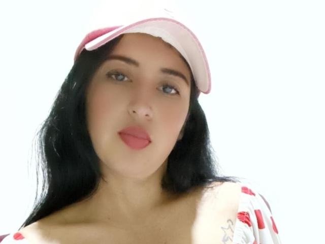 NicollJeans heeft lang zwart haar, een roze vizier, rode lippenstift en een wit topje met rode patronen, waarbij haar borsten lichtjes zichtbaar zijn. Ze kijkt naar beneden met een subtiele glimlach..