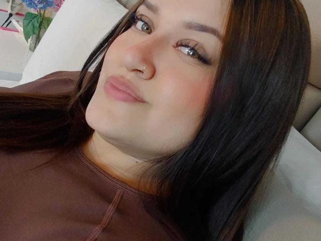 ValeriaaRose has dark brown hair, a nose ring, and pinkish lips. She's wearing a brown shirt, looking at the camera with a slight smile. Her makeup is subtle.