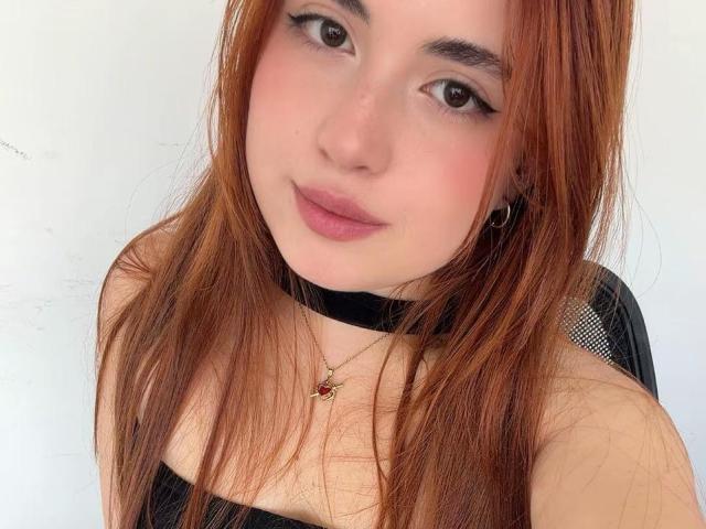 VicluX a la peau claire, les yeux marron et les cheveux roux. Elle porte un haut noir avec un collier ras du cou et un petit pendentif rouge. Elle sourit, les lèvres légèrement entrouvertes..