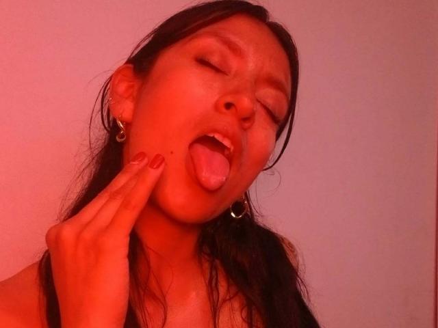 ZarayFeet has her eyes closed, tongue out, and is touching her cheek with her finger. Red lighting highlights her dark hair and hoop earrings. She's sticking out her tongue.
