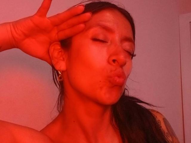 ZarayFeet with red-tinted skin, eyes closed, pouty lips, black hair in a ponytail, hand covering forehead, small hoop earrings, tattoo on shoulder, in a sensual pose.