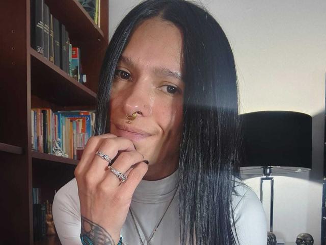 Amelie Blake, met lang zwart haar, neusring en septum piercing, lachend, hand op kin, draagt een witte blouse, meerdere ringen, tatoeage op de hand, zwarte lamp op de achtergrond..