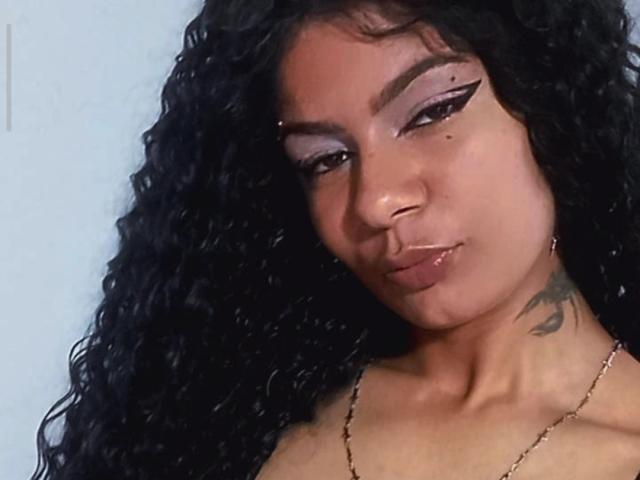 Lliizz hat lockiges, schwarzes Haar, lila Lidschatten, einen Eyeliner im "Flügel"-Stil, einen kleinen Muttermal auf ihrer rechten Wange, ein Tattoo an ihrem linken Hals und trägt eine goldene Halskette..