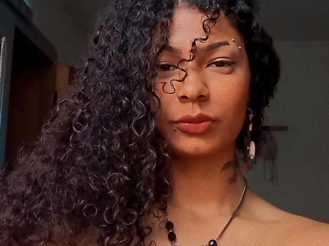 Lliizz ha i capelli neri e ricci, un piercing al naso e una collana nera. Il suo viso è serio, con gli occhi marroni, e indossa un rossetto rosso. Il suo seno non è visibile..