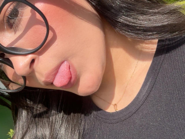 El rostro de MiiaPalmer, con la lengua afuera, usando gafas negras, con labios rosados y un ligero rubor, vestida con una camisa negra, con la luz del sol sobre su hombro, y un delgado collar dorado..