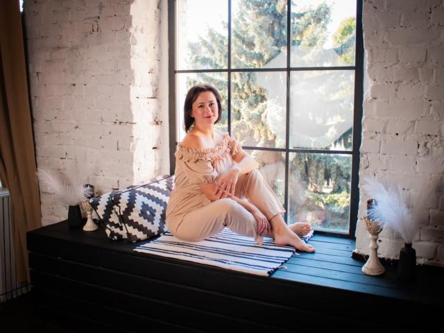 MarieSho with tits visible, sitting on a black windowsill in beige off-shoulder pants, feet on a striped rug, flanked by black and white pillows, and feather decorations.