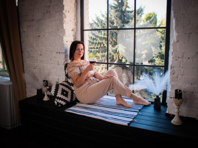 MarieSho mit Brüsten, in einem beigen Kleid, sitzend auf einem schwarzen Fensterbrett, ein Glas in der Hand, umgeben von schwarzer/weißer Dekoration und einer weißen Feder. Sonnenlicht scheint hindurch..